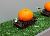 A.A.M. Bos, Orange Army (Armoured Mobile Juice Division) (2019),  die-cast toy tanks, enamel, fake plastic oranges, plastic flowers, fake grass, found shelf, detail, 16 x 120.5 x 20.5 cm.  Photo: Arekahānara